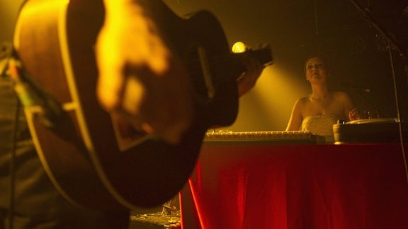 Im Vordergrund spielt eine Person Gitarre, während im Hintergrund eine Frau an einem Tisch sitzt.