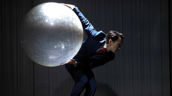 Ein Schauspieler auf einer dunklen Bühne hält einen glitzernden Gymnasitkball