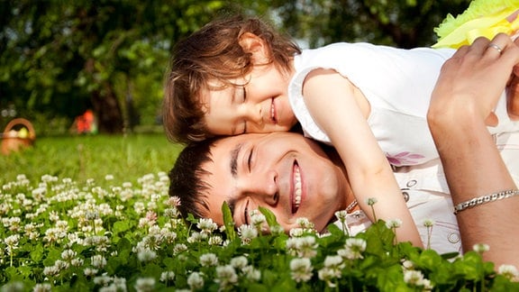 Ein Vater liegt mit seiner Tochter glücklich lachend auf einer Blumenwiese.