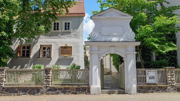 Ein Zaun mit einem steinernen Torbogen, dahinter ein Garten und ein Haus