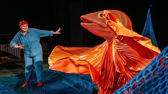 Eine Person mit blauer Kleidung und roter Mütze steht auf der Bühne. Eine große Puppe eines Goldfischs fliegt auf sie zu.