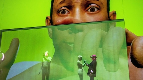 Vor einem grünen Hintergrund ist das überdimensionale Gesicht eines Mannes zu sehen, im Vordergrund stehen drei Personen