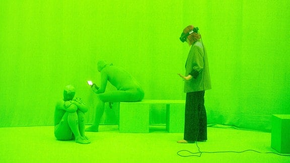 Vor grünem Hintergrund steht eine Frau mit VR-Brille, daneben sitzen zwei Personen in Ganzkörperanzügen.  