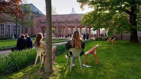 Personen sitzen auf Designerstühlen auf der Wiese des Grassimuseums, dessen markantes Gebäude sich mit seinen Torbögen im Hintergrund abzeichnet.