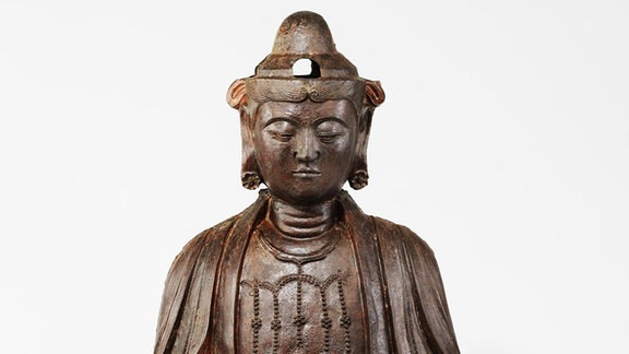 Eine gegossene Eisenskulptur (buddhistische Kultplastik): Der altertümlich, flache Körper ist vom Gewand her eher einem Buddha zuzuordnen, dazu ein weicher modellierter Bodhisattvakopf