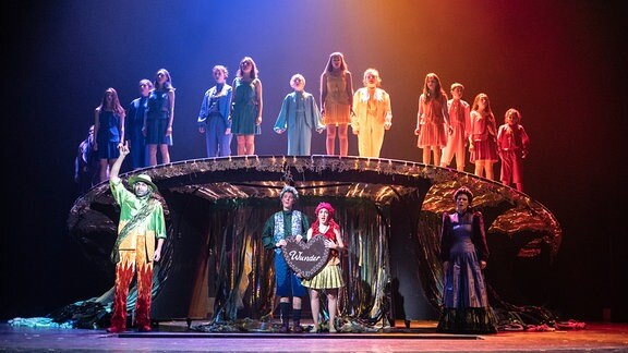 Ein Chor in Regenbogenfarben steht auf einem Podest, davor mehrere weitere Personen in bunten Kleidern.