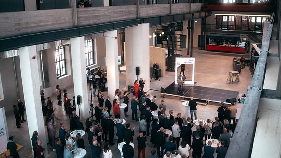 In einer großen Halle stehen viele Menschen vor einer Bühne an Stehtischen