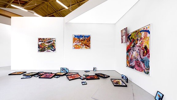Blick in einen weißen Ausstellungsraum mit farbstarken Bildern an den Wänden und auf dem Boden liegend