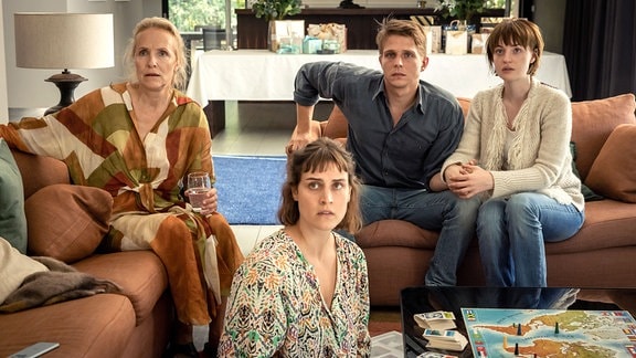 Szene aus der Serie "Haus aus Glas": eine vierköpfige Familie sitzt um einen Couchtisch herum und schaut erschrocken