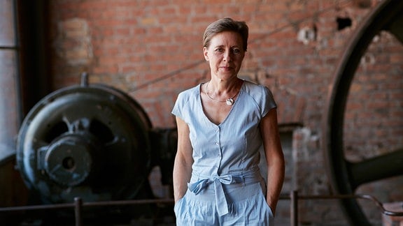 Annette Schlünz, eine Frau in einem hellblauen Einteiler steht vor einer Backsteinmauer