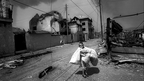 Schwarz-Weiß-Foto: Eine Person trägt riesige Säcke über eine unbefestigte Straße zwischen Zement-Häusern.