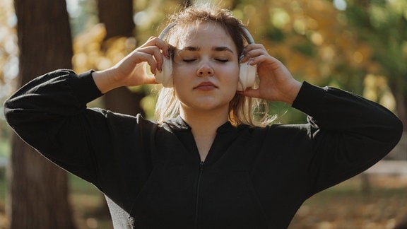 Junge Frau mit geschlossenen Augen, die über drahtlose Kopfhörer im Park Musik hört.