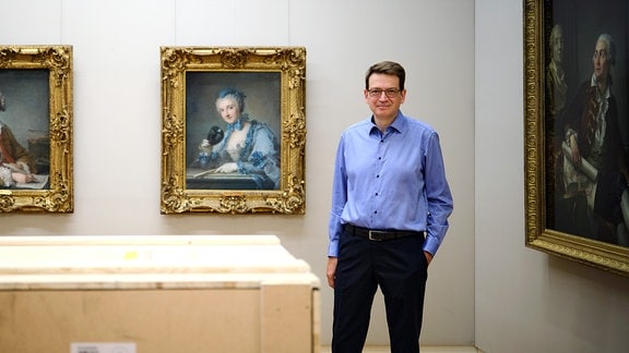 Holger Jacob-Friesen: Ein Mann mit blauem Hemd steht lässig neben riesigen Kisten in einem Ausstellungsraum mit klassischen Gemälden an den Wänden.