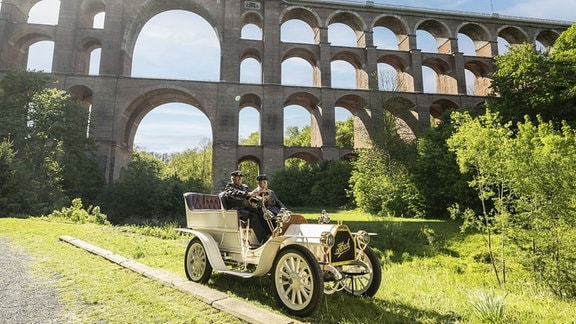 Ein historisches Automobil mit zwei Personen vor einer Bogenbrücke.