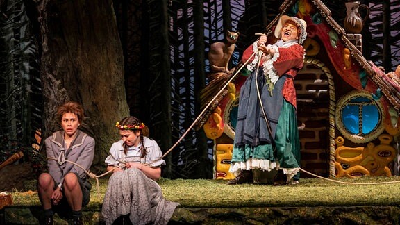 Szene aus dem Märchentheaterstück "Hänsel und Gretel": Zwei Menschen sitzen am Bühnenrand. Eine Hexe zieht sie mit einem Seil zu ihrem Lebkuchenhaus.