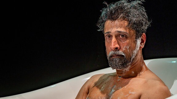 Ein Mann sitzt in einer Badewanne, Schaum hängt in seinem Gesicht und er schaut traurig ins Leere.