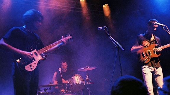 Auf einer bunt erleuchteten Bühne stehen zwei Menschen mit Gitarren an Standmikros und einer sitzt am Schlagzeug.