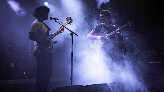 Zwei Personen mit GItarren stehen sich auf einer nebligen Bühne gegenüber.