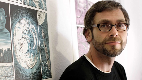 Ein Mann mit Fünf-Tage-Bart und Brille steht vor einer Wand mit Zeichnungen.