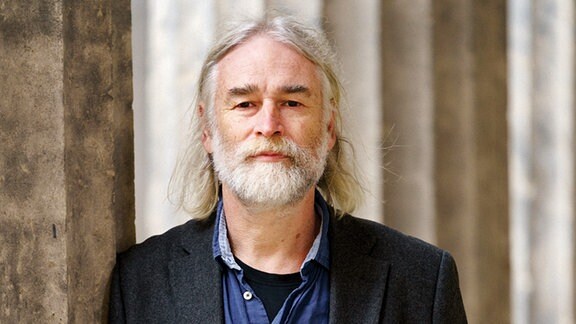 Autor Jens Wonneberger: ein Mann mit grauem Haar und Bart lehnt an einer Säule und blickt in die Kamera