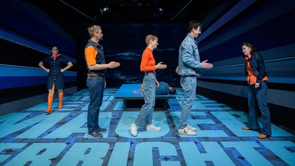 Drei Menschen stehen mit angewinkelten Armen in einer Reihe hintereinander auf einer blauen Bühne.