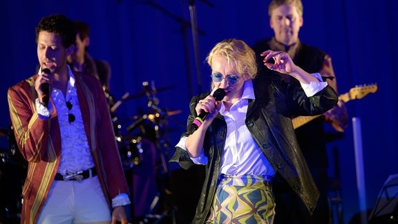Eine Frau mit kurzen blonden Haaren, Lederjacke, runder Sonnenbrille und bunter Hose singt in ein Mikrofon. Im Hintergrund sind weitere Teile einer Band zu sehen.