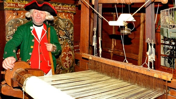 Ein Mann mit Kostüm des Räuberhauptmanns Karasek steht an einem Webstuhl. Er trägt einen Dreispitz, eine grüne Jacke und rote Weste.