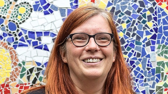 Eine Frau mit roten Haaren und Brille steht vor einer Wand mit bunten Mosaiksteinen und lacht in die Kamera.