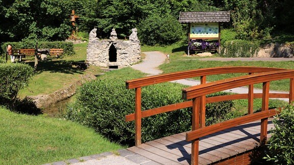 Eine schmale Brücke aus Holz, im Hintergrund Bänke, eine überdachte Hinweistafel, eine gemauerte Grotte, Wiese und Bäume. 