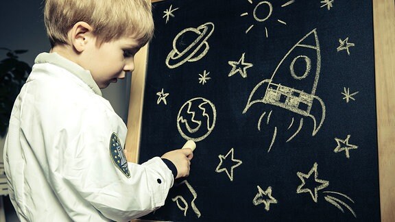 Ein Junge malt Planeten, Sterne und eine Rakete auf eine Tafel