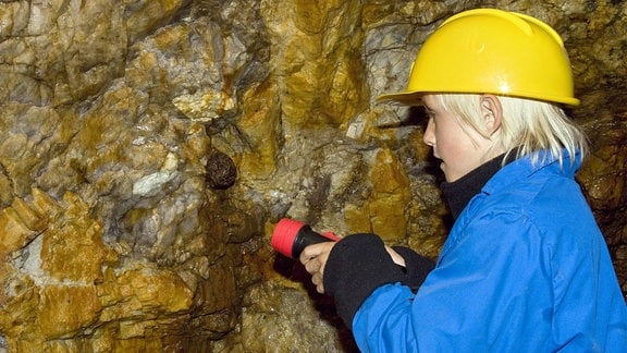 Kind besichtigt ein Bergwerk unter Tage, entdeckt mit Taschenlampe Mineralien an den Wänden des Stollens.