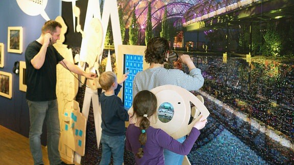 Ein Mann, eine Frau und zwei Kinder stehen in einem Museum vor einer Wand mit dem "Messe-M" der Leipziger Messe. 