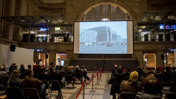 Eine Kinofilm-Vorführung im Bahnhof von Leipzig: eine Leinwand, vor der Publikum sitzt
