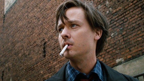 Ein junger Mann mit einer Zigarette im Mund vor einer Backsteinwand. 