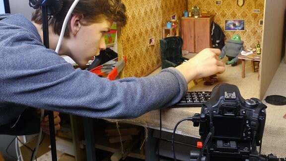 Ein junger Mann bei Arbeiten an einem Stop-Motion-Film, eine Fotokamera ist auf ein kleines Modellzimmer mit Figuren gerichtet.