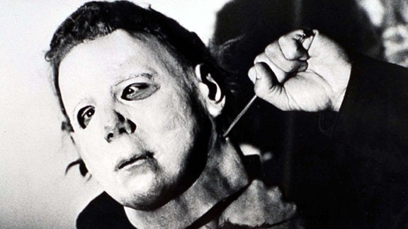 Schwarz-Weiß Szene aus dem Film "Halloween". Mann mit weißer Maske sticht sich einen spitzen Gegenstand in den Hals. 