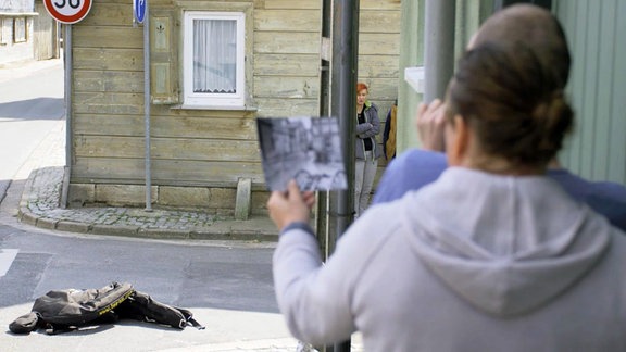 Im Vordergrund ist eine Person von hinten zu sehen, die ein schwarz-weiß-Foto in der Hand hält, das man nicht erkennt. Sie Person blickt auf eine Straßenecke, an der eine lebensgroße Puppe liegt.