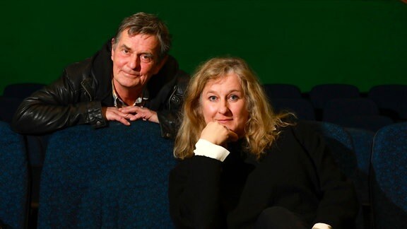 Ein Mann mit grauen Haaren und einer schwarzen Lederjacke und eine Frau mit langen blonden Haaren sitzen auf dunkelblauen Kinosesseln. 