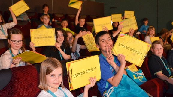 Kinder sitzen in einem Kinosaal, sie halten Klemmbretter mit einem Papier in die Höhe, auf dem obersten gelben Papier steht in Großbuchstaben "Streng geheim".