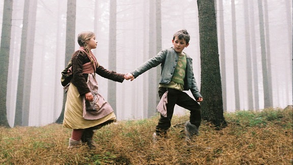 Filmszene aus "Hänsel und Gretel", die Kinder halten sich an der Hand und laufen durch den Wald.