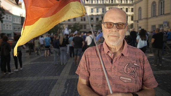 Thomas Pischel steht mit einer Deutschlandfahne in der Hand auf einem Platz, auf dem eine Demonstration stattfindet.