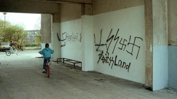 Ein Kind fährt auf einem Fahrrad durch einen Hausdurchgang in einem Plattenbauviertel, an die Wand wurden Hakenkreuze und SS-Symbole geschmiert.