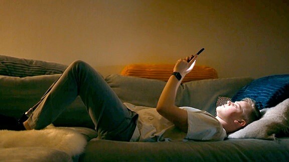 Alex, ein Protagonist der Serie "Cyborgs of Instagram", liegt auf dem Sofa und blickt auf sein Handy.
