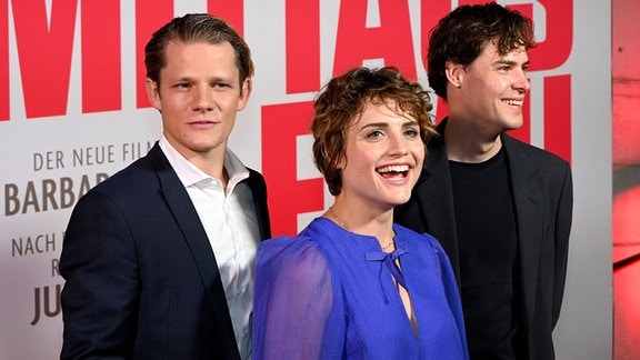 Zwei junge Männer und eine junge Frau stehen vor dem Werbeplakat für eine Film.