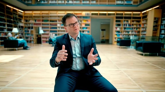 Ein Mann mit Brille und Anzug sitzt in einem großen lichten Raum mit Bücherwänden