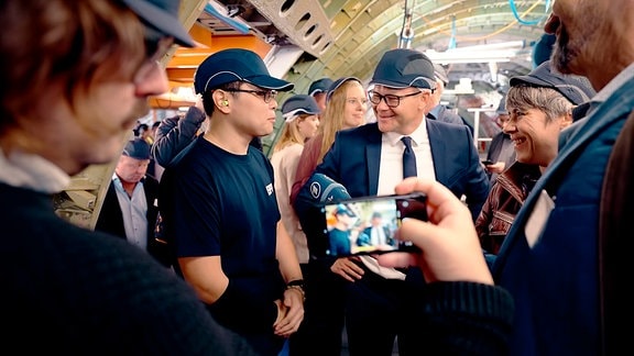 Ein Mann mit Brille und im Anzug spricht lächelnd mit einem jungen Mann mit Käppi und dunkelblauer Werkskleidung umringt von anderen Menschen in einer Industriehalle