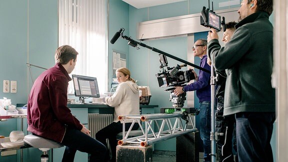Eine blonde Frau mit weißem Pulli sitzt an einem PC, neben ihr ein Mann in Jeans und roter Sweatjacke. Hinter ihnen sind Mitglieder eines Filmteams zu sehen, die Kamera, Tonangel und Kontrollbildschirm halten.