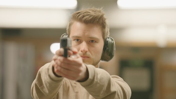 Erik Koszuta, ein junger Mann mit hellen, kurzen Haaren, etwas Bart und einem hellbraunen Pullover trägt einen Gehörschutz und hält in seinen ausgestreckten Armen eine Pistole. 