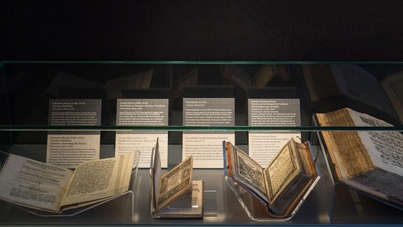 Vier aufgeschlagene, historische Gesangsbücher liegen in einer beleuchteten Vitrine. Daneben sind Informationstafeln zu sehen. 