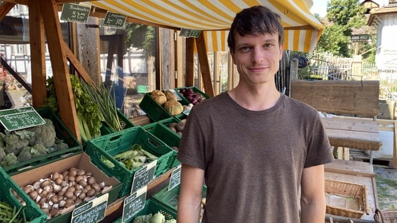 Ein junger Mann in braunem T-Shirt steht unter einer Markise vor grünen Kisten voller Gemüse 